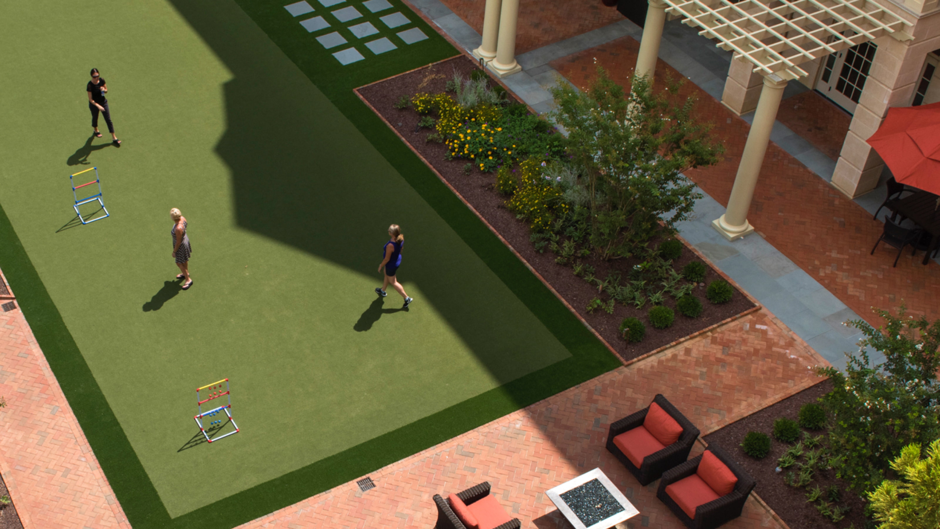 Aerial view of a courtyard with a green lawn, brick walkways, a seating area with red cushions, and a small garden with yellow flowers surrounded by shrubs. Three people are walking across the lawn.